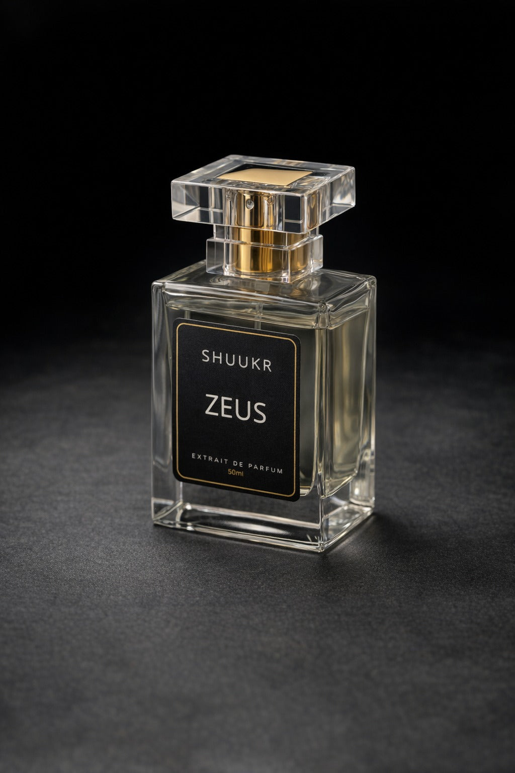 A clear glass perfume bottle with a square cap and a black front label reading 'SHUUKR ZEUS', photographed on a dark textured surface against a black background.